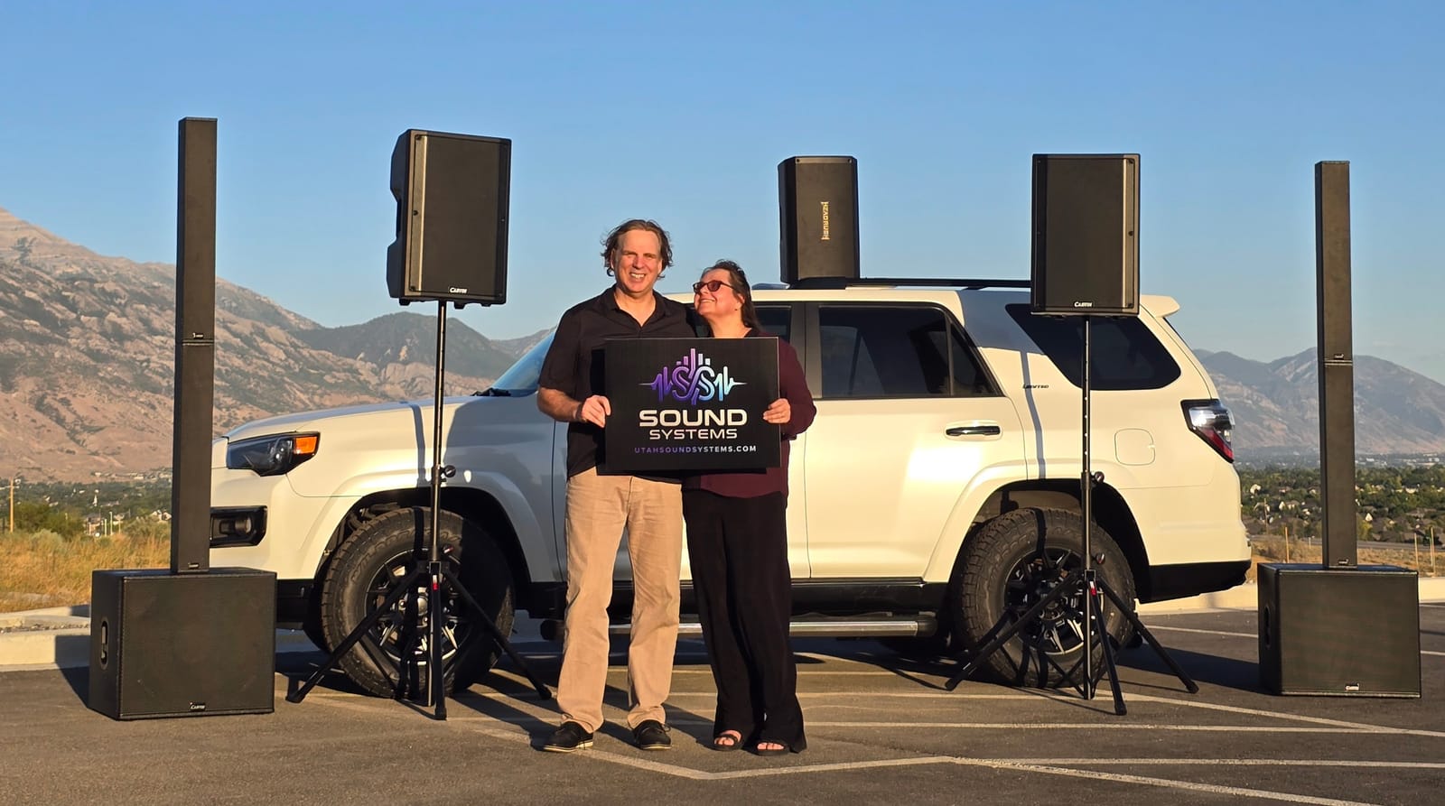 Two people holding a Sound Systems sign in front of the branded company SUV, flanked by column array speakers, powered speakers on stands, and subwoofers, with Utah mountains behind