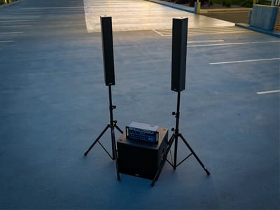 Two column array speakers raised on tripod stands either side of a mixer and subwoofer at dusk