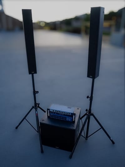 A powered 18-inch subwoofer on the ground with a column array and mixer stacked above it, staged at dusk