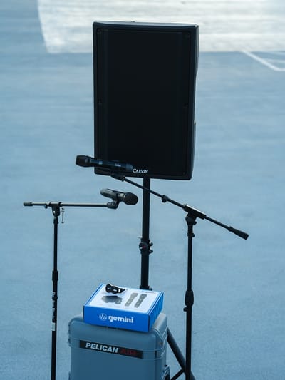 A Carvin speaker on a stand, two microphone stands, and a boxed Gemini wireless microphone system resting on a Pelican Air case