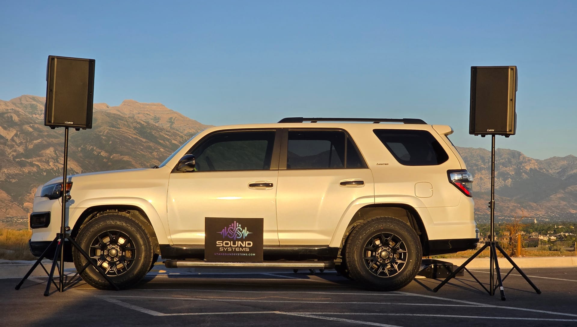 Two Carvin QXSYS15A speakers raised on tripod stands at either end of the branded Sound Systems SUV, with the Wasatch range behind them at golden hour