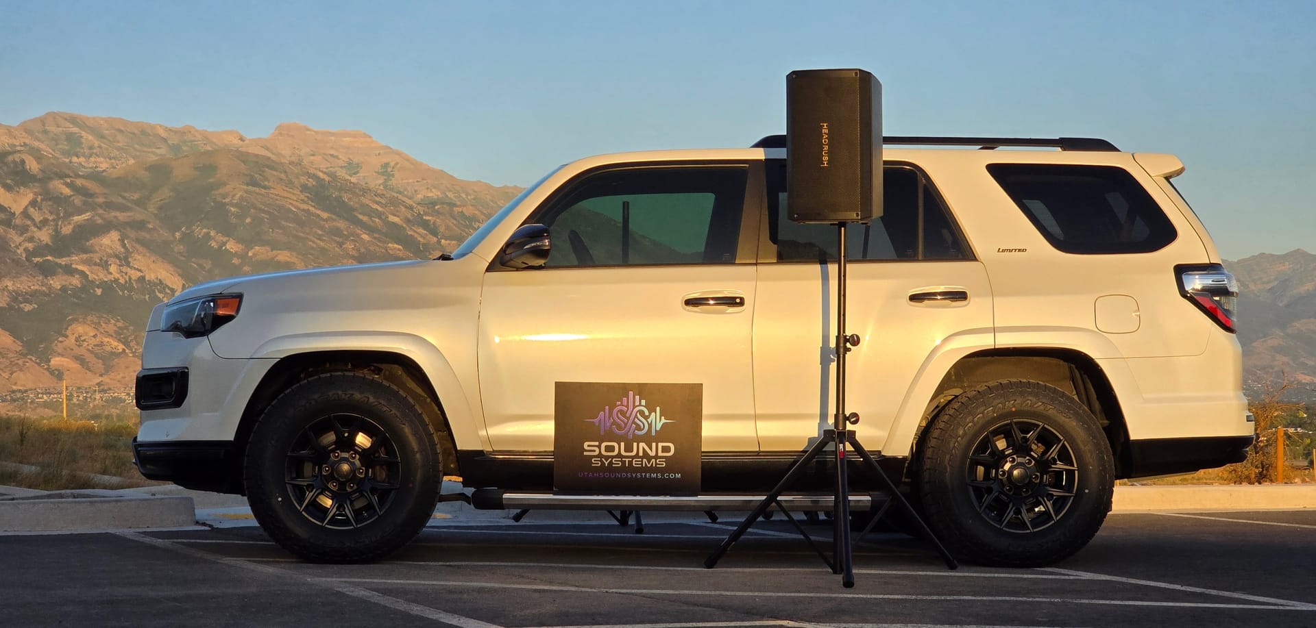The branded Sound Systems SUV parked at golden hour with a HeadRush powered speaker raised on a tripod stand beside it and the Wasatch mountains behind