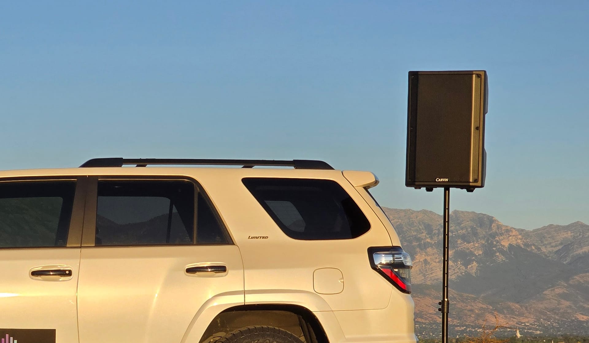 A Carvin QXSYS15A powered speaker raised on a tripod stand beside the Sound Systems SUV, the Wasatch range behind it at golden hour
